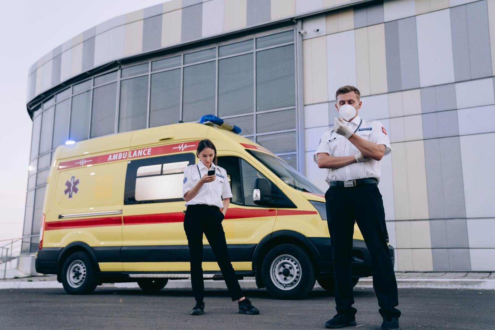 Two paramedics in uniform standing by an ambulance outside a modern building, ready to respond. - 2016 QHP landscape data