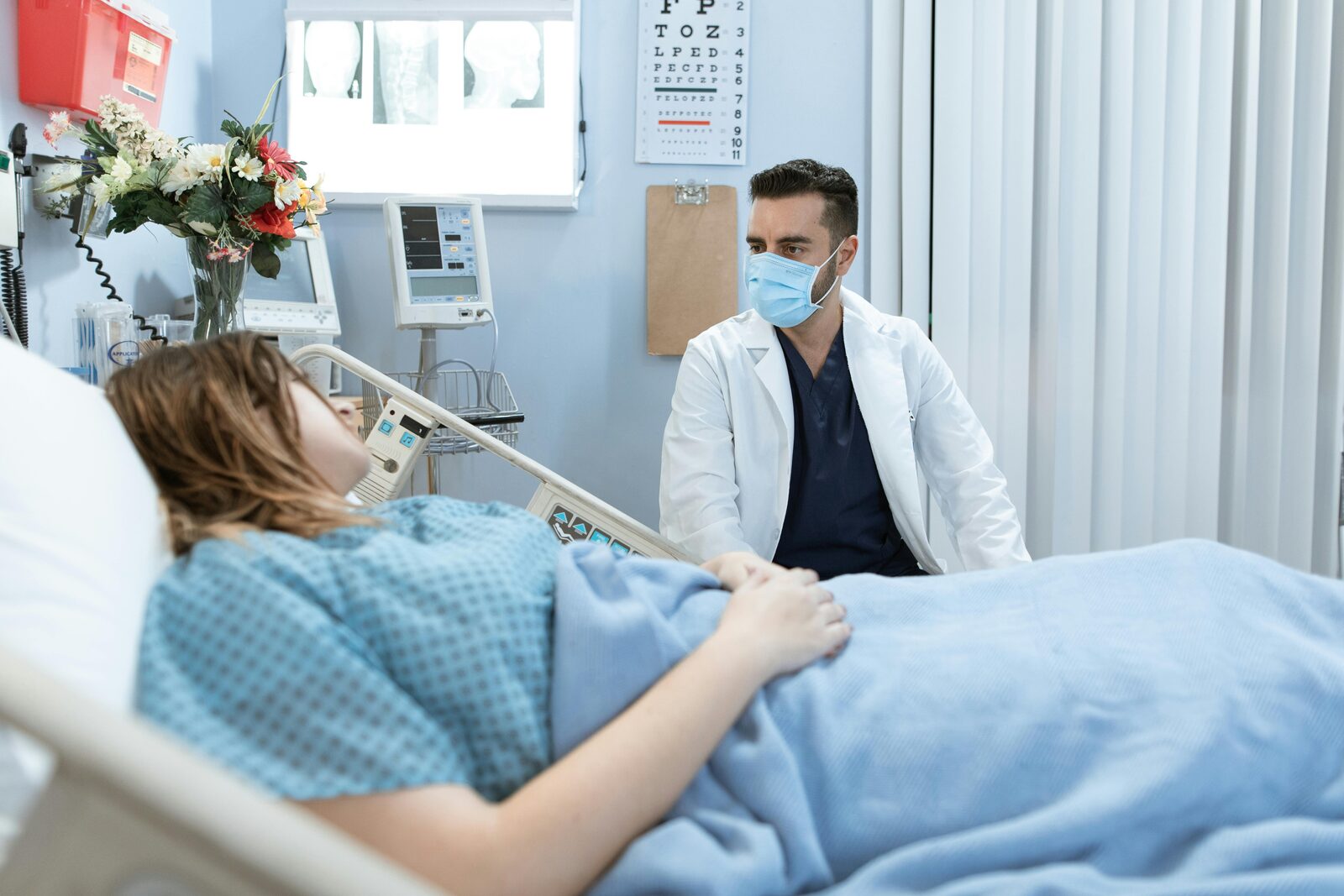 Short-Term Medical — A doctor wearing a face mask sits beside a patient in a hospital bed, providing care and consultation, representing short-term medical coverage.