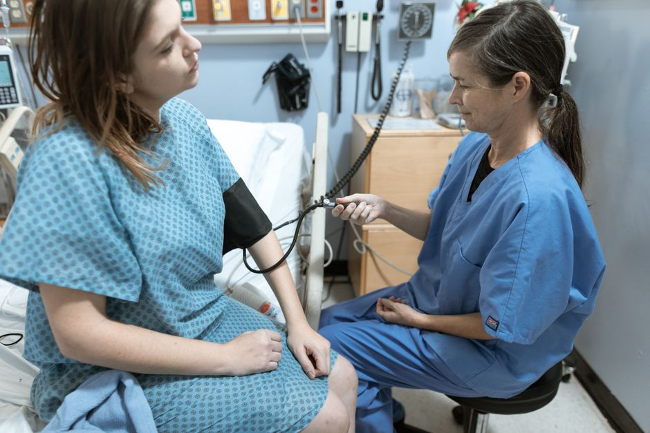 A nurse in blue scrubs checks the blood pressure of a female patient in a hospital room. health insurance