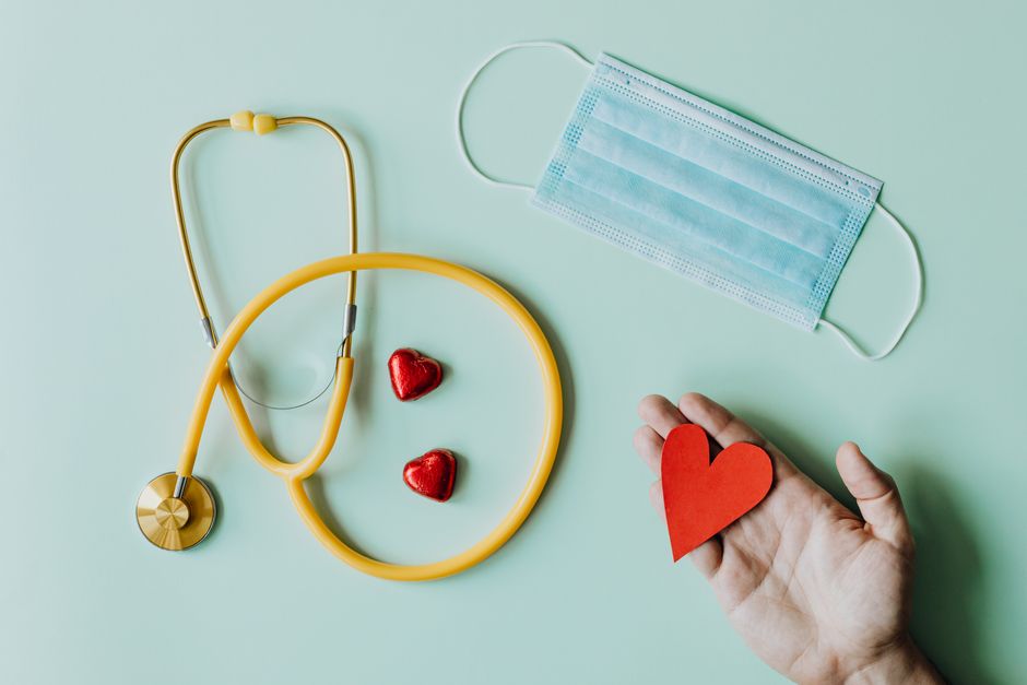 Top view of crop anonymous person hand with red paper heart on table with stethoscope and medical mask for coronavirus prevention health insurance