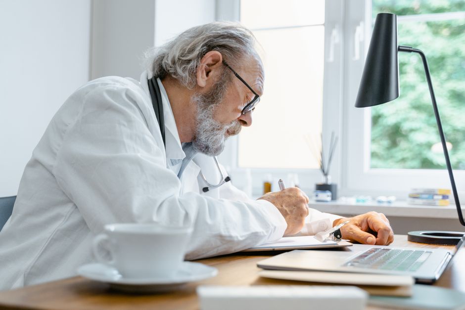 Elderly doctor writing notes at his desk with a focus on work in a well-lit office. health insurance