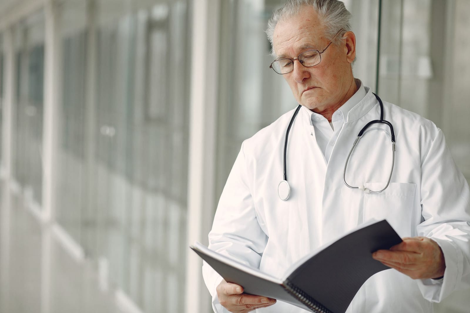 Crop concentrated professional senior male physician in white robe with stethoscope standing in modern hospital hallway and reading medical case records in daylight health insurance