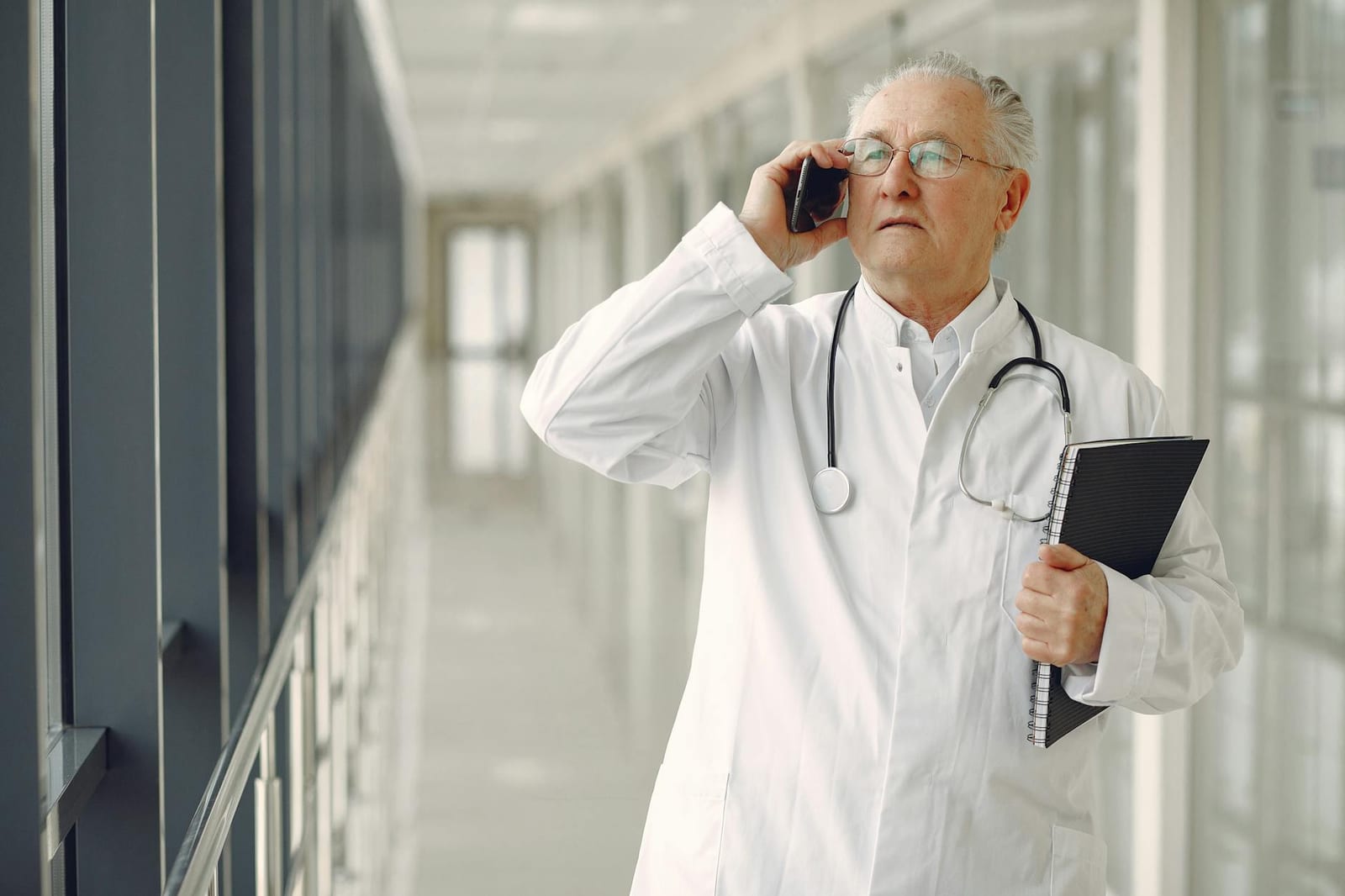 Serious mature male doctor in medical uniform standing with stethoscope and notebook while talking on smartphone in clinic corridor health insurance