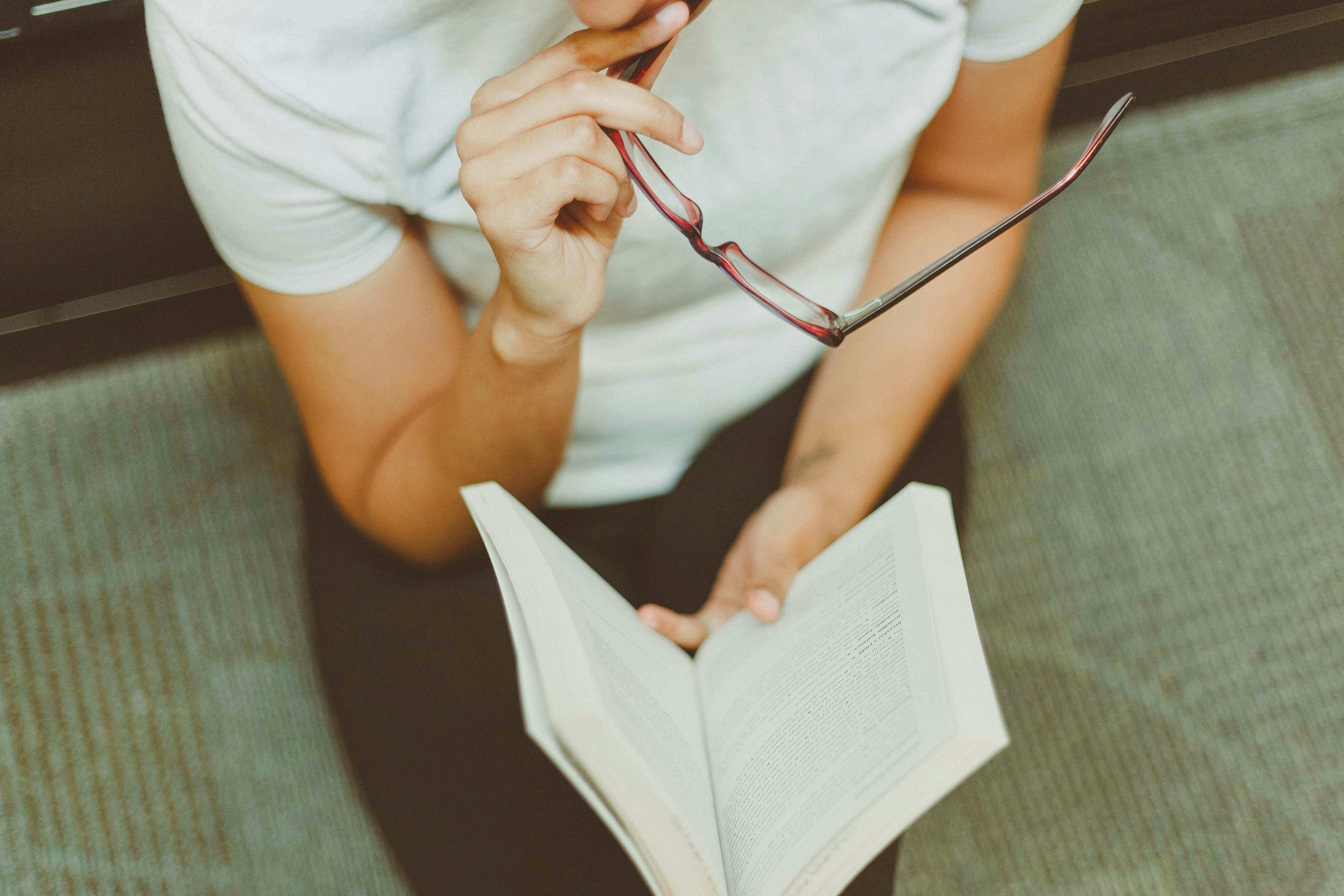 Health Insurance Learning Center: Guides & Articles Person sitting on a couch holding red-framed glasses while reading an open book, representing the health insurance learning center and educational guides.